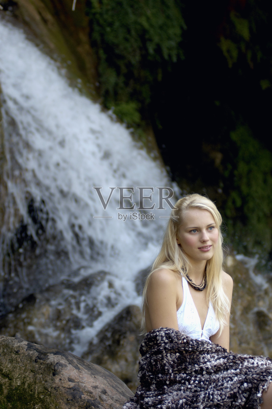 年轻女子坐在瀑布边,裹着毯子照片摄影图片