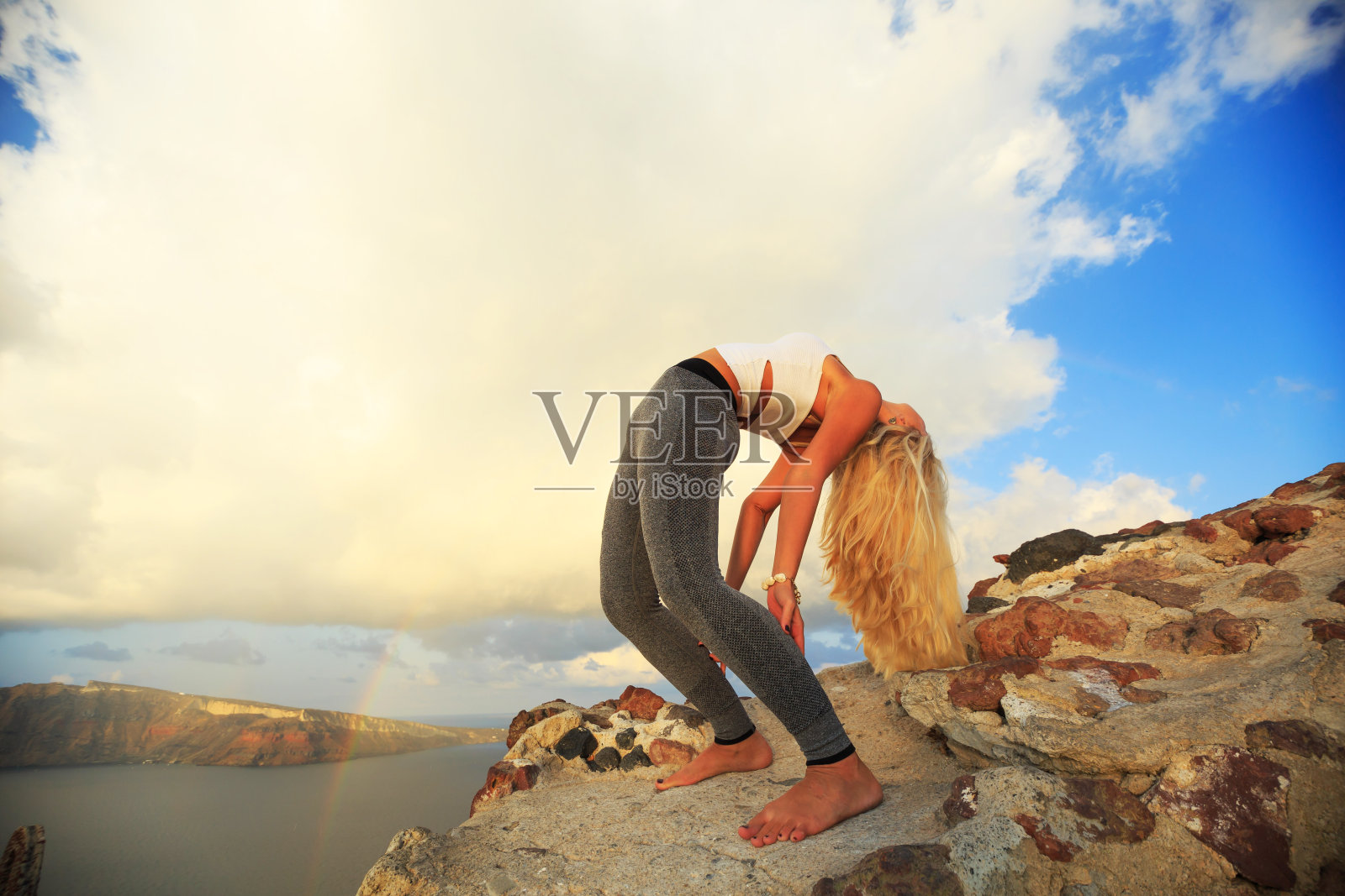 金发的年轻女子在希腊圣托里尼的岩石练习瑜伽照片摄影图片