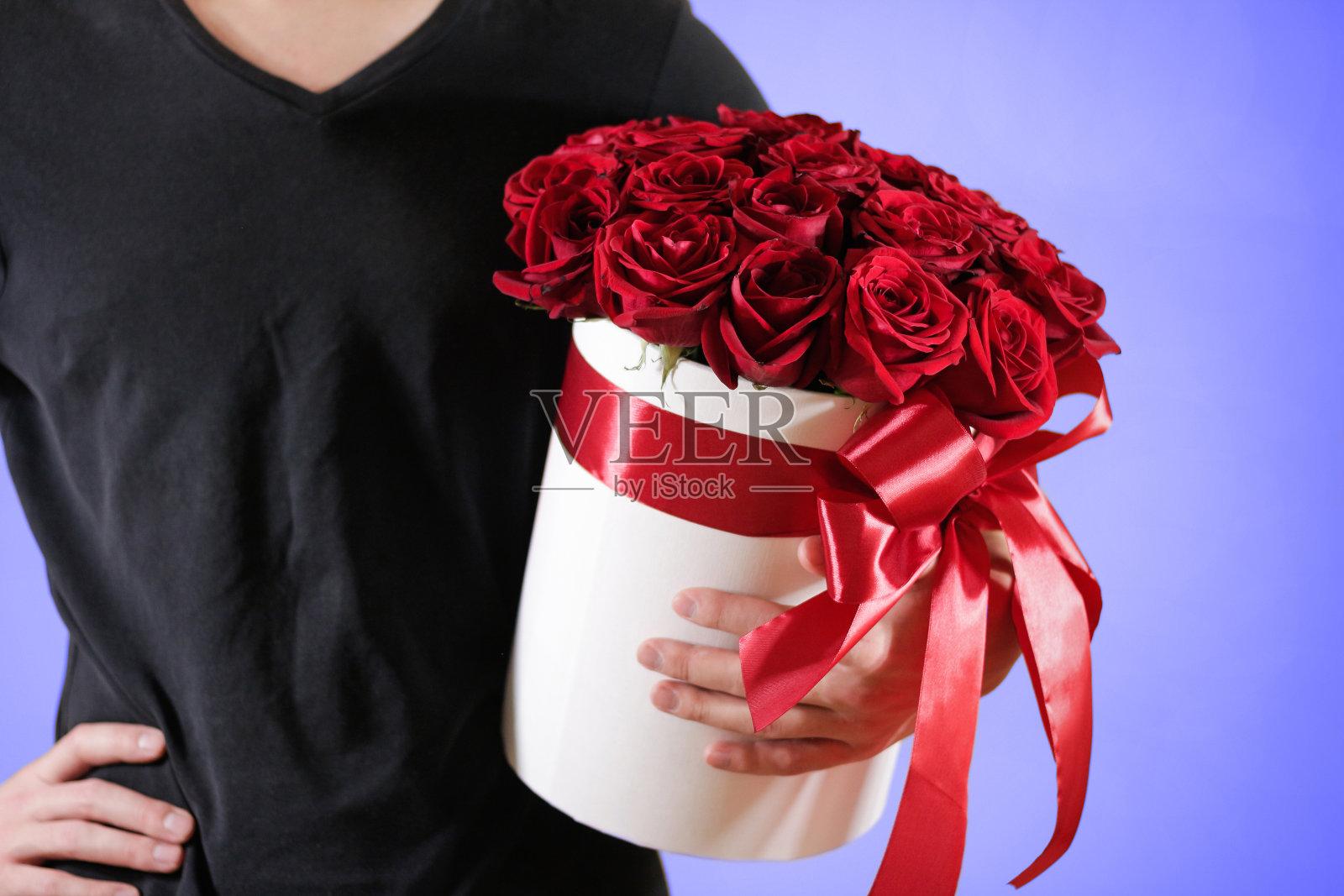 男子穿着黑色t恤,手里拿着21朵红色玫瑰的富丽礼品花束。白色帽盒中的鲜花组成。用红丝带和蝴蝶结系好。照片摄影图片