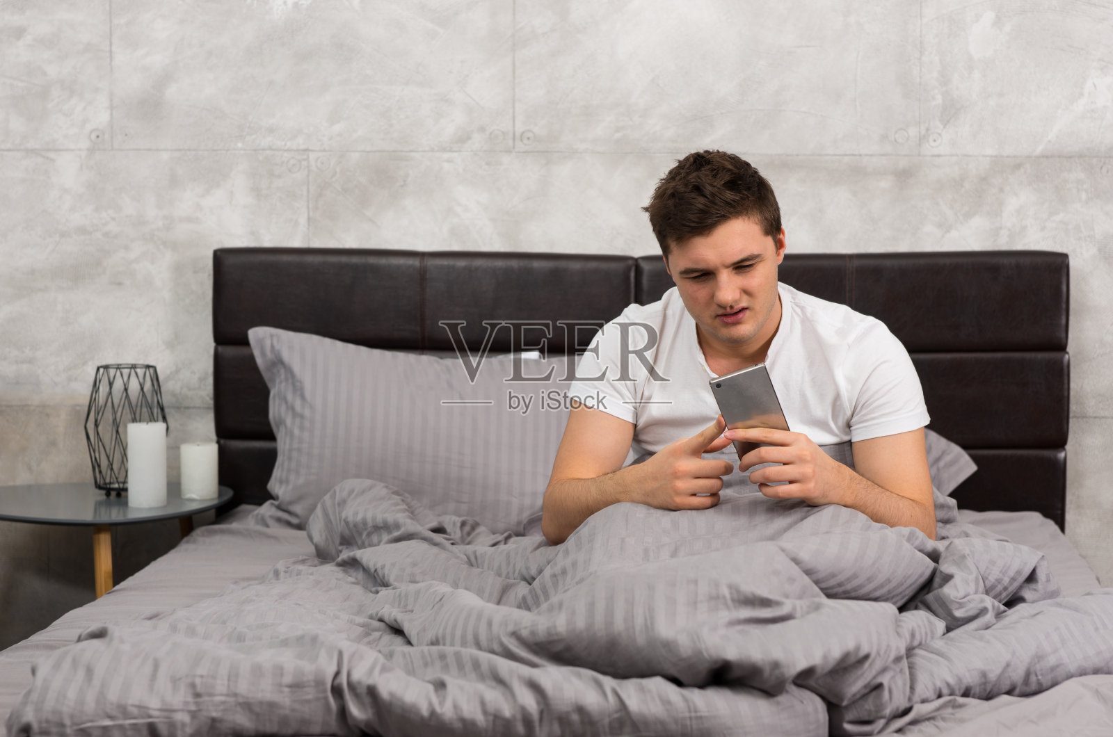 一个年轻人醒了过来,坐在时髦的灰色床上看他的手机照片摄影图片