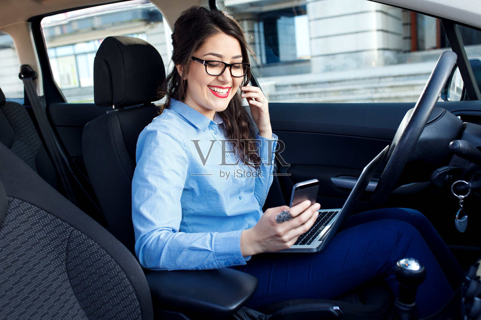 年轻漂亮的女商人坐在车里用笔记本电脑看她的智能手机。照片摄影图片