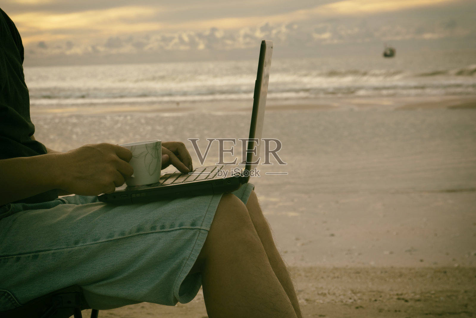 一个在海滩上拿着杯子用笔记本电脑的男人。照片摄影图片