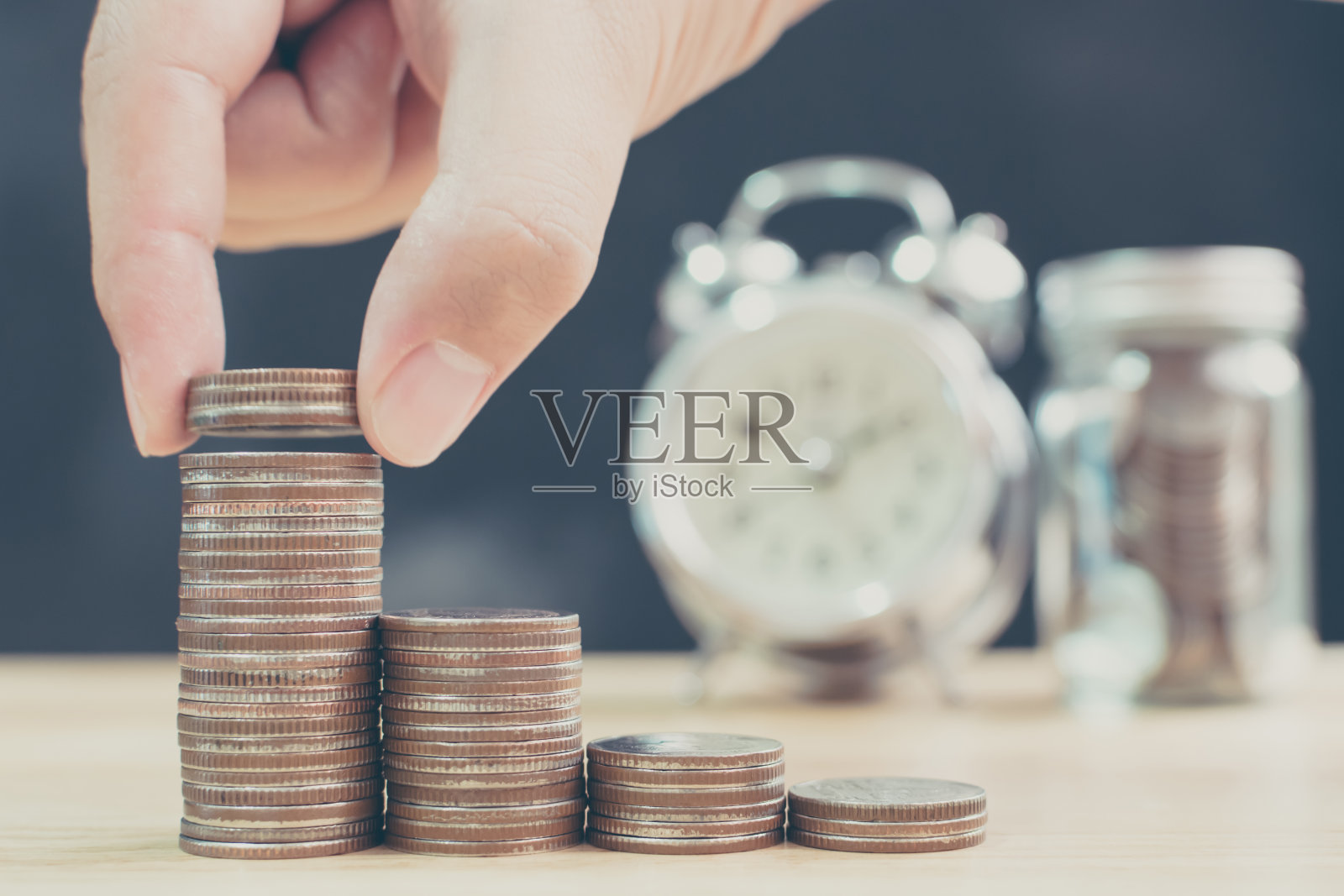 男女手把硬币堆叠起来,以模糊时钟和存钱罐为背景,理财和投资理念照片摄影图片