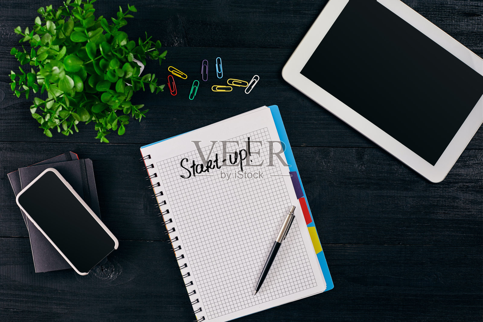 俯视图的打开笔记本上写着启动的题词。绿色花朵,平板,彩色回形针,智能,钢笔和笔记本木制背景照片摄影图片