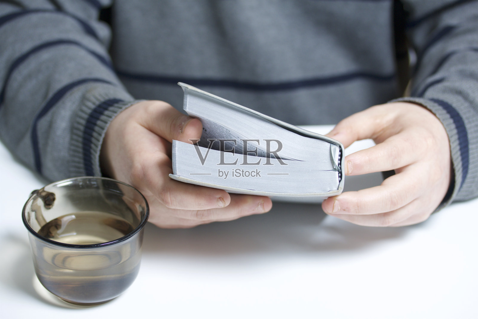 一个年轻人拿着银片坐在桌子旁读圣经。旁边放着一个装满水的杯子。照片摄影图片