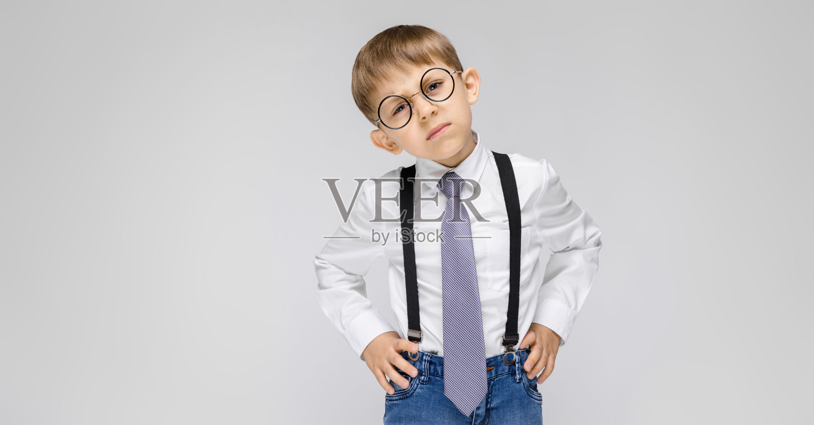 灰色背景上站着一个可爱的男孩,他穿着白衬衫、背带、领带和浅色牛仔裤。戴眼镜的男孩把头歪向身边照片摄影图片