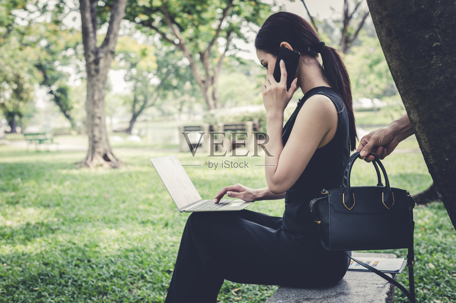 小偷的手在女子身后捡起包,她坐在花园里和朋友用智能手机聊天。罪犯、恶棍、土匪的概念照片摄影图片