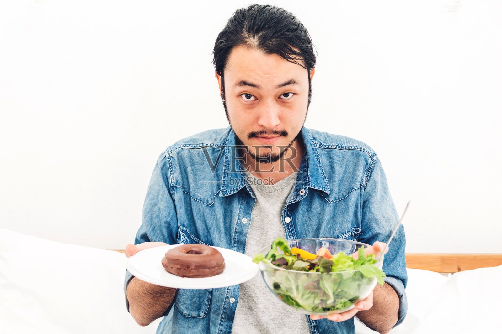 男人抱着和选择健康的沙拉和卡路里炸弹巧克力甜甜圈在床上在家。健康饮食和垃圾食品的概念照片摄影图片