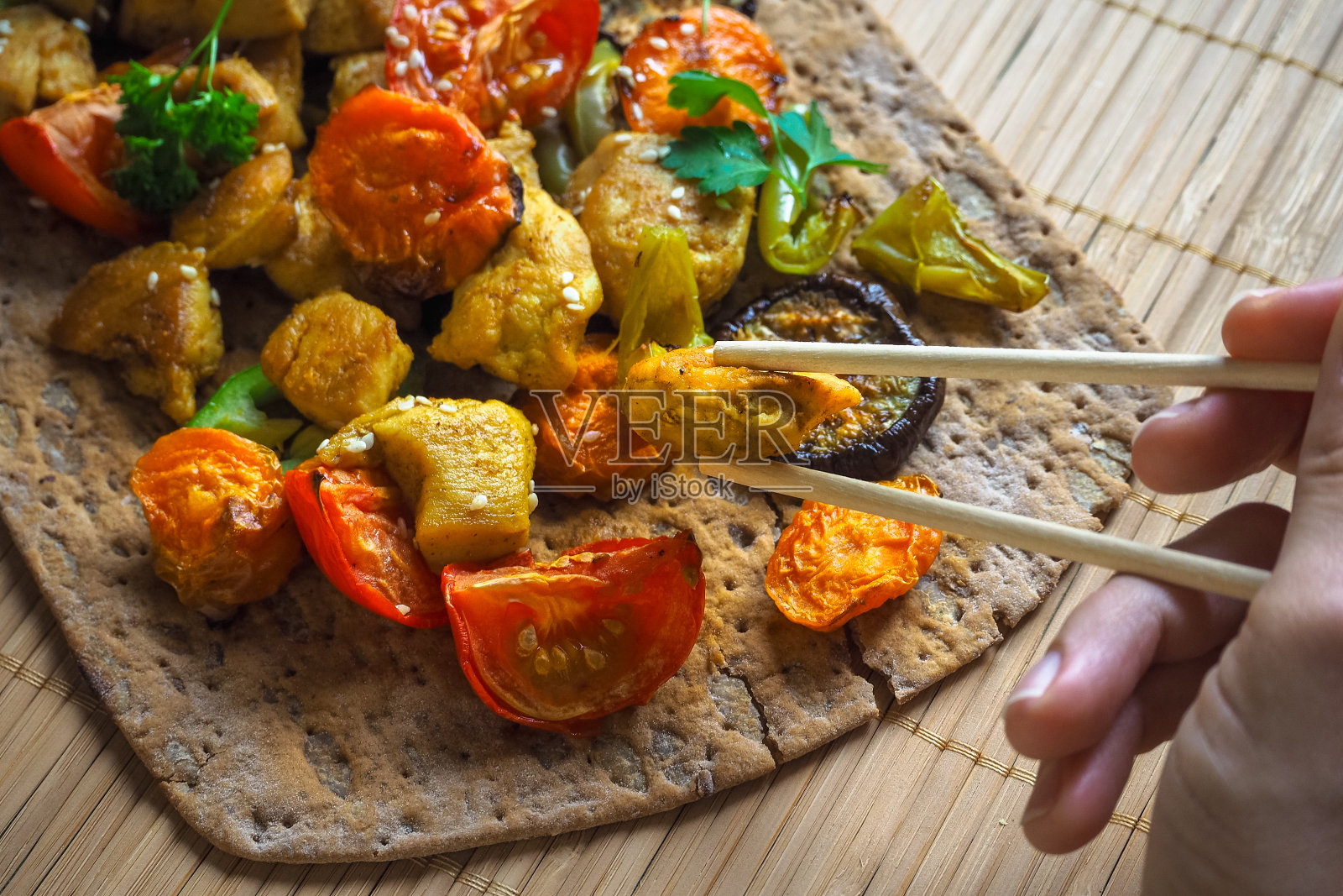用筷子夹着一块鸡肉的手。烤制的蔬菜配咖喱鸡片。照片摄影图片
