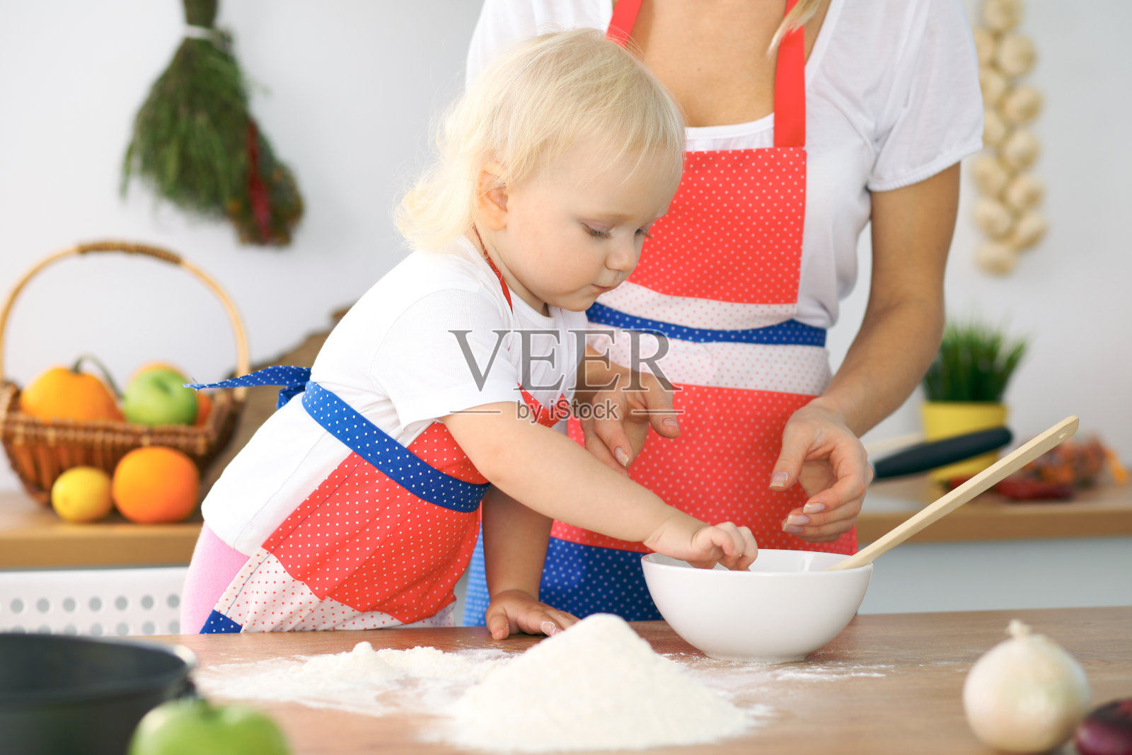 小女孩和她金发碧眼的妈妈穿着红色围裙在厨房里揉面团,边玩边笑。自制糕点,面包,比萨饼或烤饼干照片摄影图片