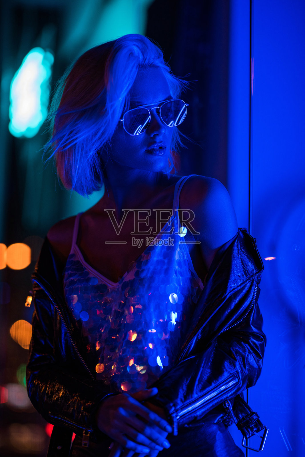 美丽的年轻女子在蓝色的灯光下穿着光滑的背心和太阳镜在街道上照片摄影图片