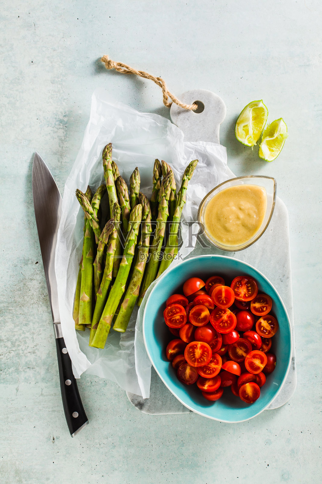 新鲜健康的食材包括绿芦笋、圣女果、芝麻酱和酸橙酱,放在大理石切菜板上和厨刀上照片摄影图片