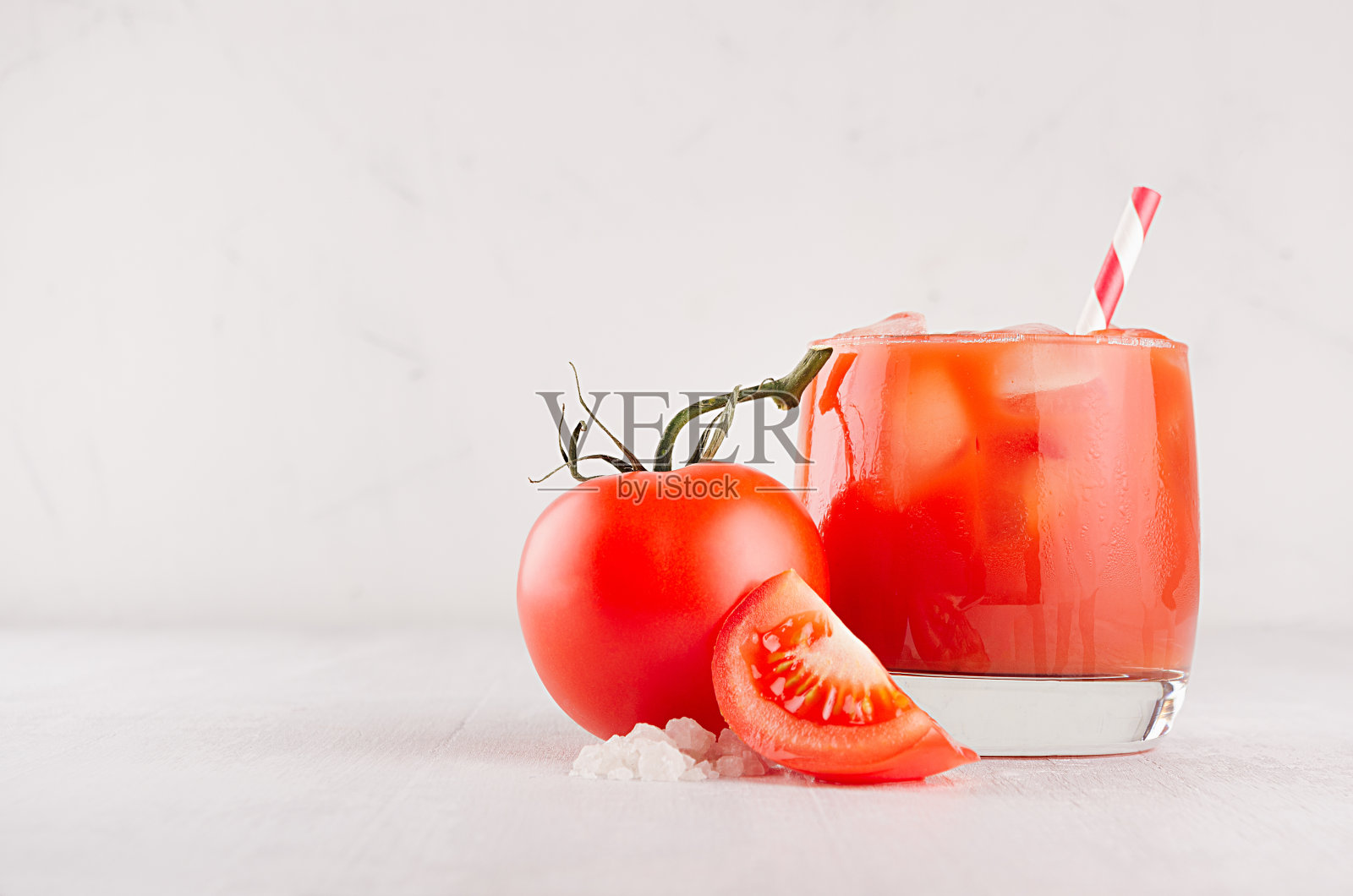 鲜亮的冷红色番茄鸡尾酒血腥玛丽优雅的玻璃杯,有光泽的番茄,盐,吸管,冰块和一块白色木板。照片摄影图片
