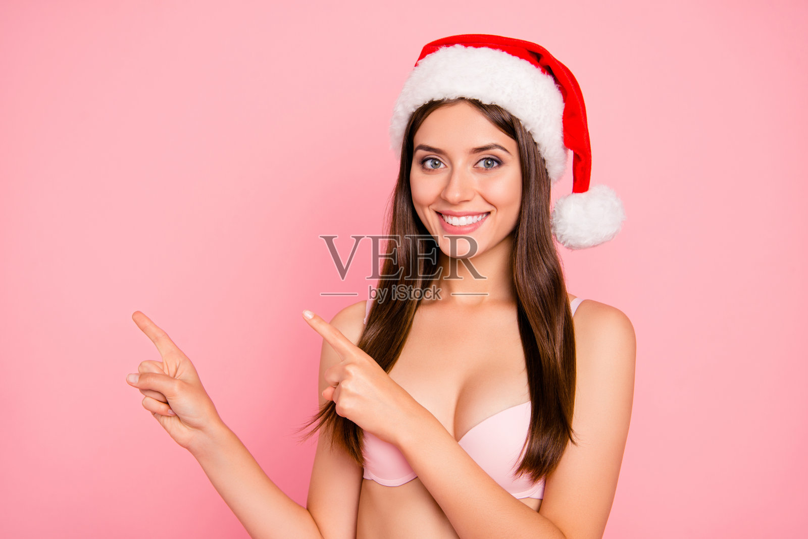 漂亮迷人的迷人的积极愉快的华丽甜美的直发女士在内衣和帽子,指着两个手指一边的肖像,复制空间,孤立在粉色柔和的背景照片摄影图片