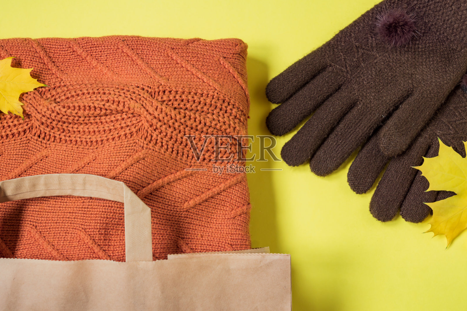 橙色女人温暖的毛衣在纸工艺包装和棕色手套在黄色的背景与秋天枫叶装饰。服装促销和黑色星期五概念。前视图。照片摄影图片