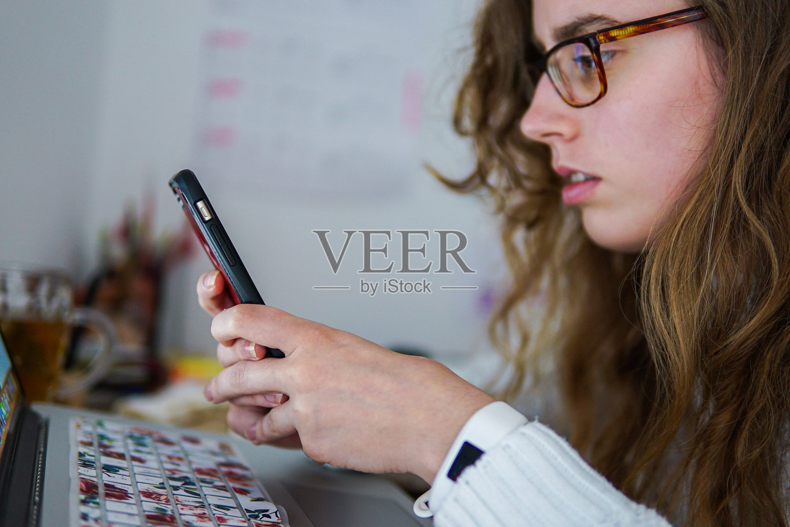 女孩看着手机在她的电脑前,桌子上摆满了东西。照片摄影图片