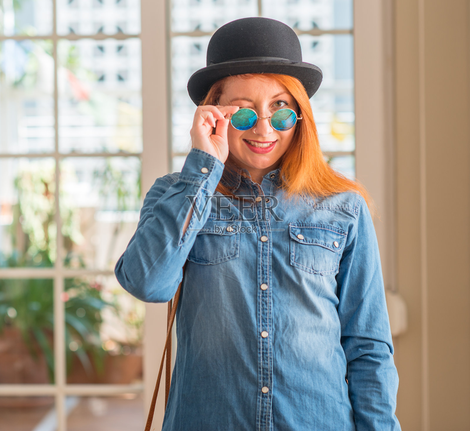 时尚的红发女人戴着圆顶礼帽和太阳镜看着镜头,性感的表情,快乐的脸。照片摄影图片