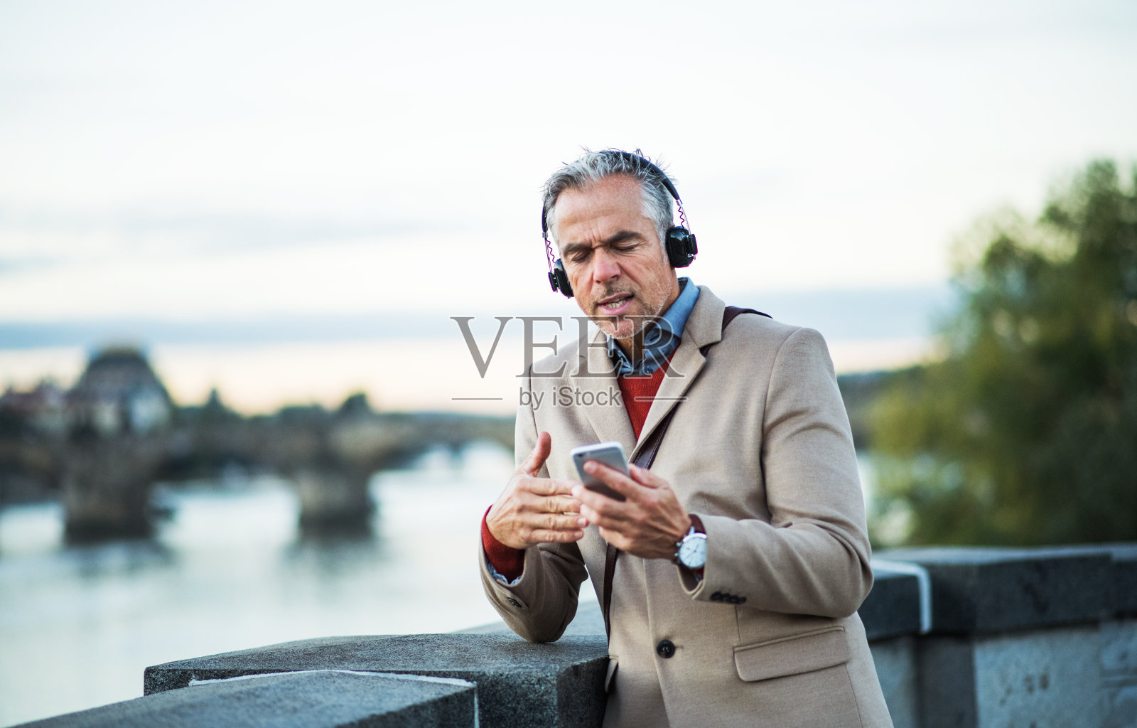 成熟的商人戴着耳机和智能手机站在河边唱歌。照片摄影图片