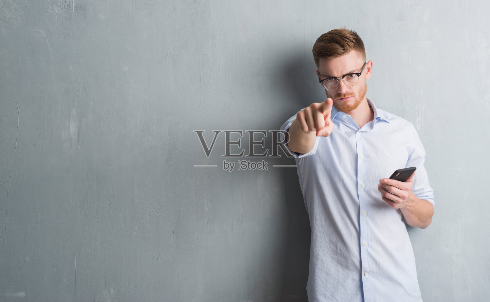 年轻的红发男子越过灰色的垃圾墙,用智能手机用手指指着相机和你,手势,正面自信的手势发送信息照片摄影图片