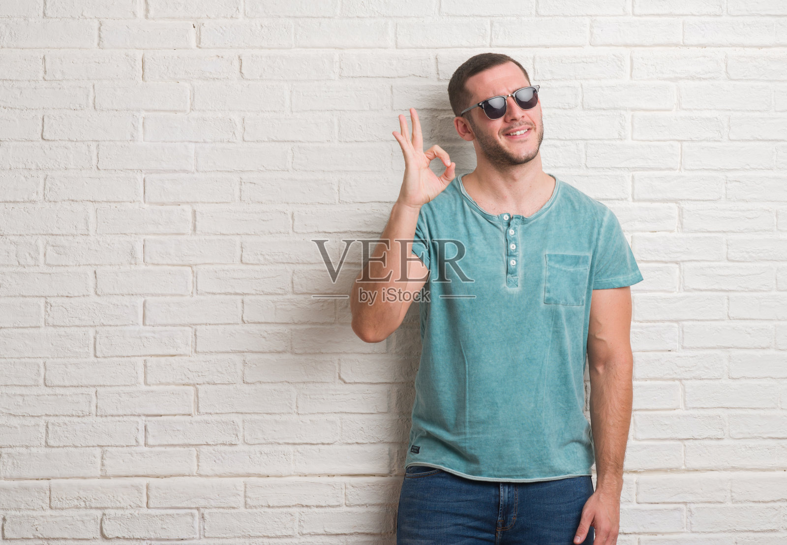 一个年轻的白人男子戴着墨镜站在白砖墙前用手指做ok手势,很好的象征照片摄影图片
