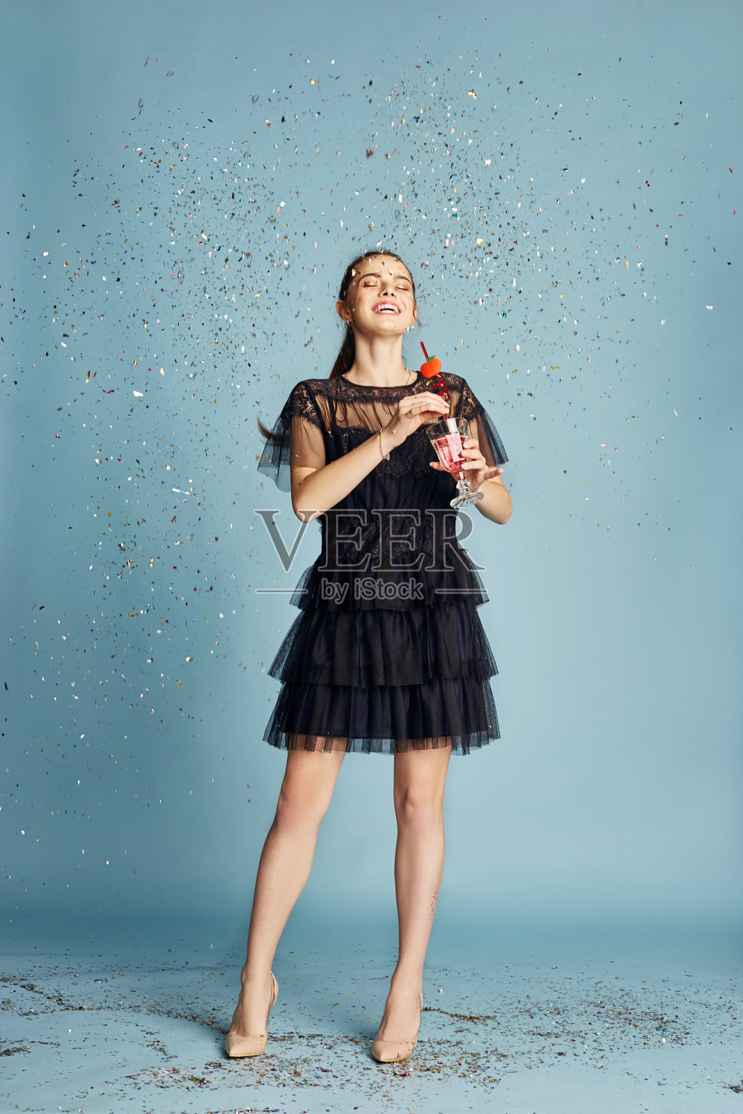 美丽的女人庆祝生日派对,在飞舞的五彩纸屑下开心地笑着吃蛋糕。女孩摆姿势和微笑在蓝色的背景,愉快的情绪,微笑和笑声照片摄影图片