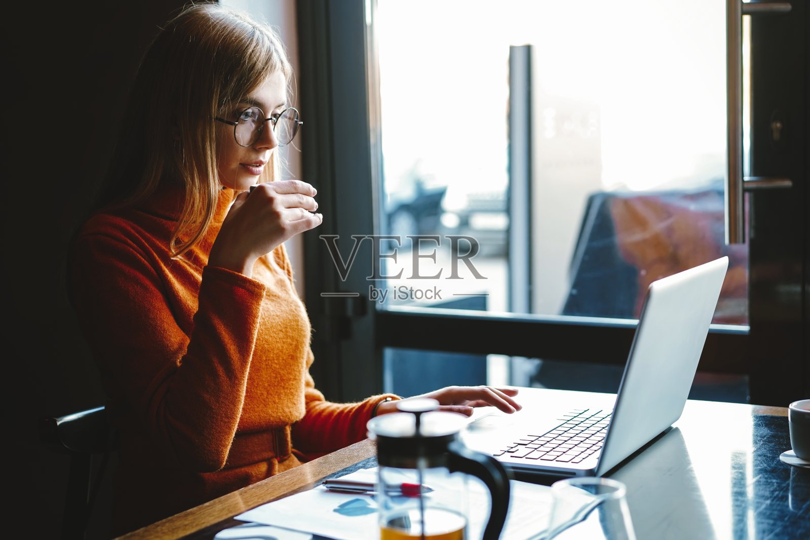 集中在工作上。自信的年轻女子,穿着时髦的休闲装,坐在创意办公室或咖啡馆靠窗的地方用笔记本电脑工作。照片摄影图片