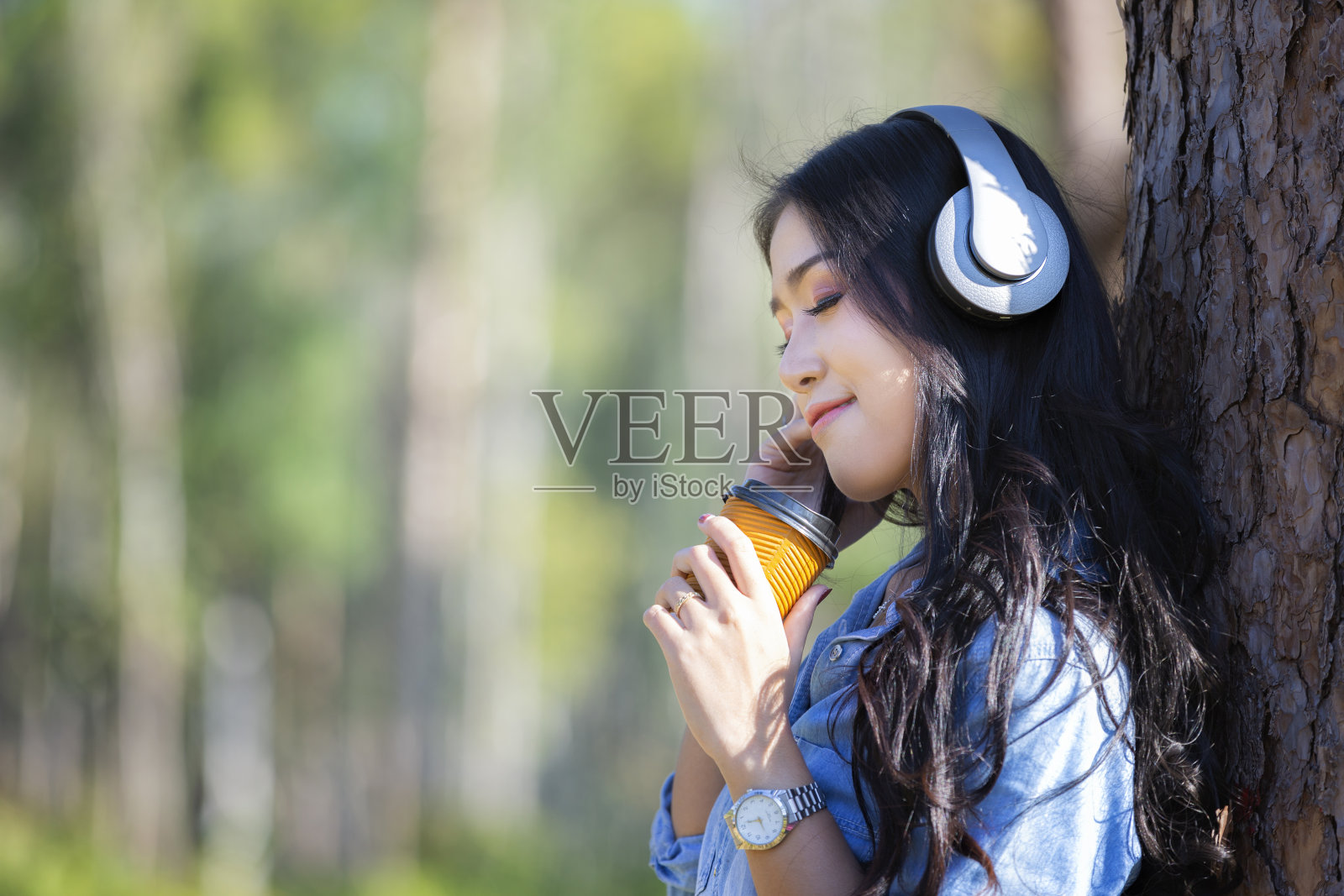 年轻女子戴着现代耳机在松林里一边喝着咖啡一边在线交流。照片摄影图片