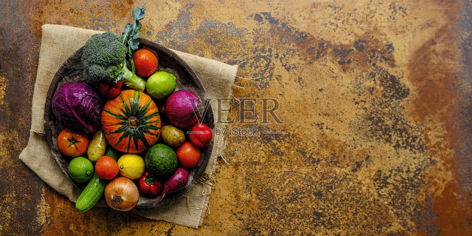 许多色彩鲜明的沙拉蔬菜和柠檬和酸橙,坐在一个圆形的,旧的,木制蔬菜碗上,麻袋材料,在抽象的陶土乡村背景与大气的照明。照片摄影图片