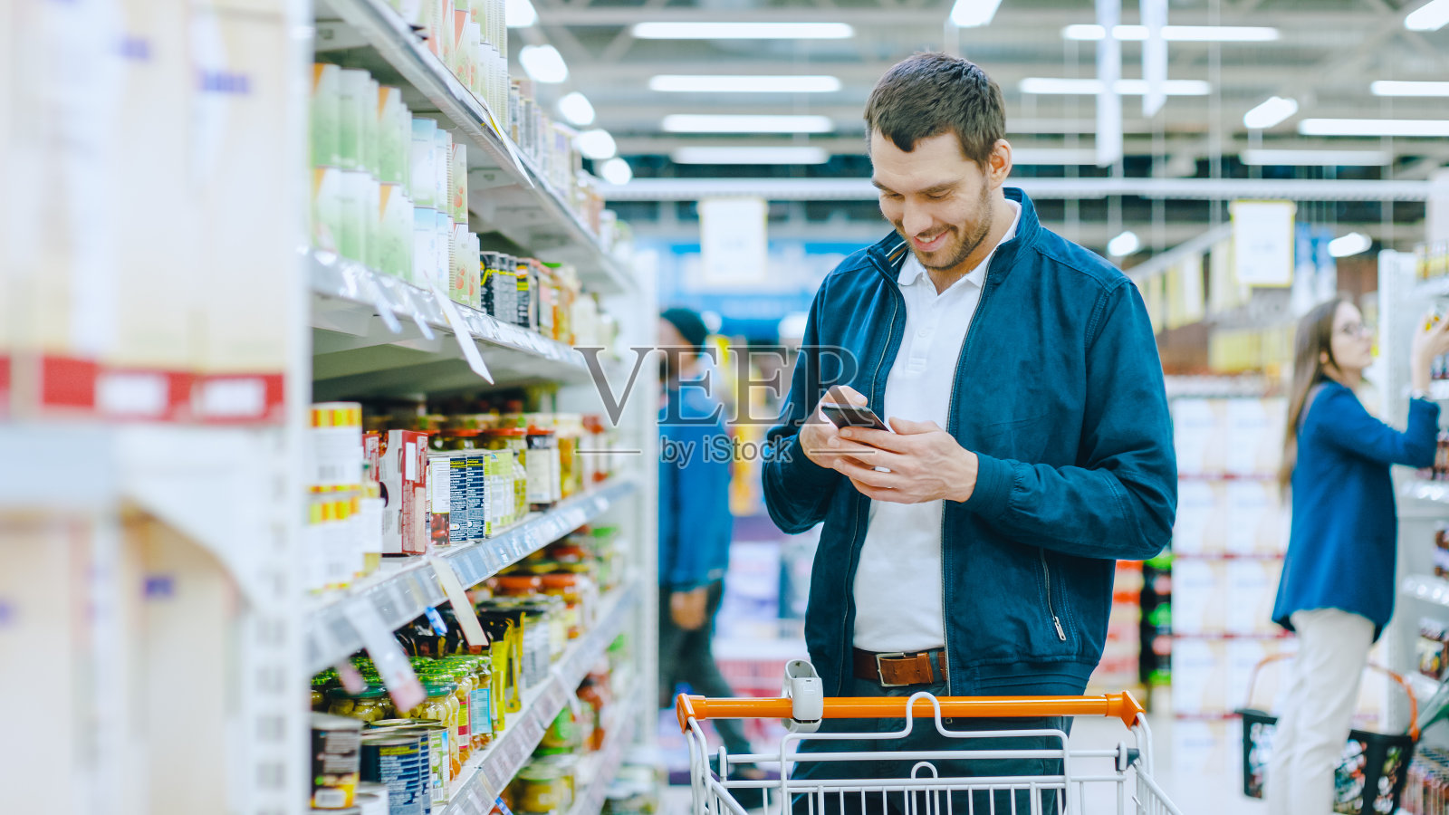 在超市:英俊的男人使用智能手机和在罐头食品区。他在寻找最好的健康产品照片摄影图片