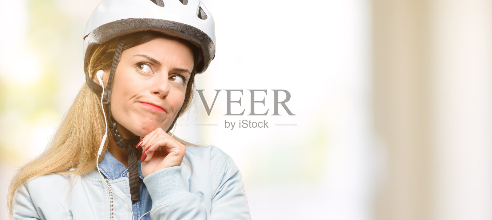 年轻女子戴着自行车头盔和耳机,一边思考一边抬头,表达着怀疑和疑惑照片摄影图片