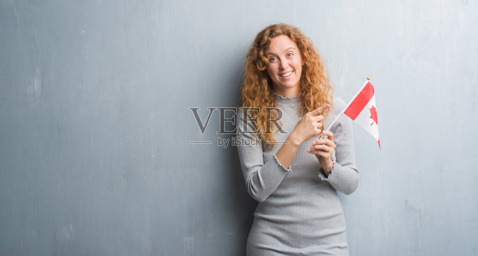 年轻的红头发的女人在灰色的墙举着加拿大国旗非常高兴的手指和手指照片摄影图片