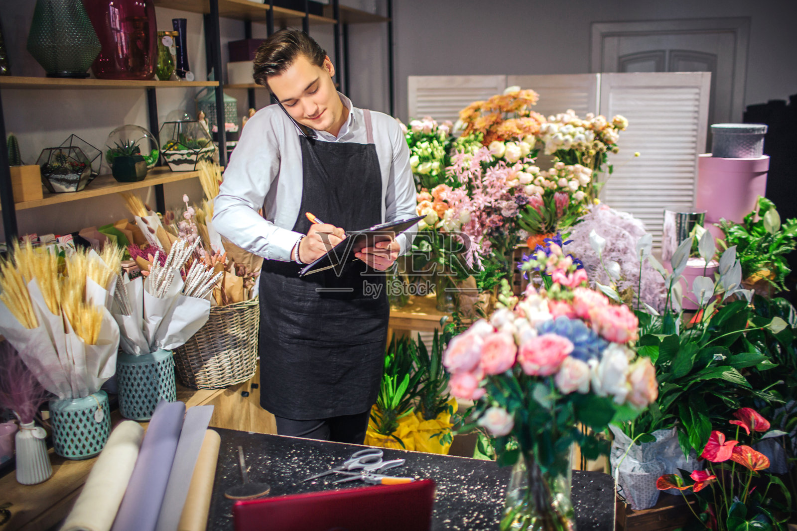 图片的年轻男性花商谈电话和白色下来的黑色板。他有笔记本电脑,花瓶和桌上的纸卷。他是忙碌的。照片摄影图片