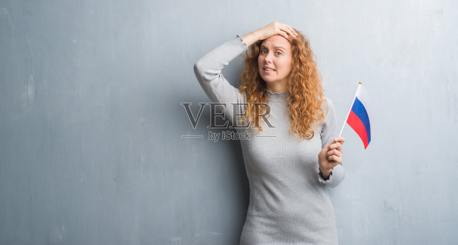 年轻红发女子站在灰色墙壁前拿着俄罗斯国旗,手扶着头,感到有压力,震惊、羞愧和惊讶,愤怒和沮丧,害怕和不安照片摄影图片