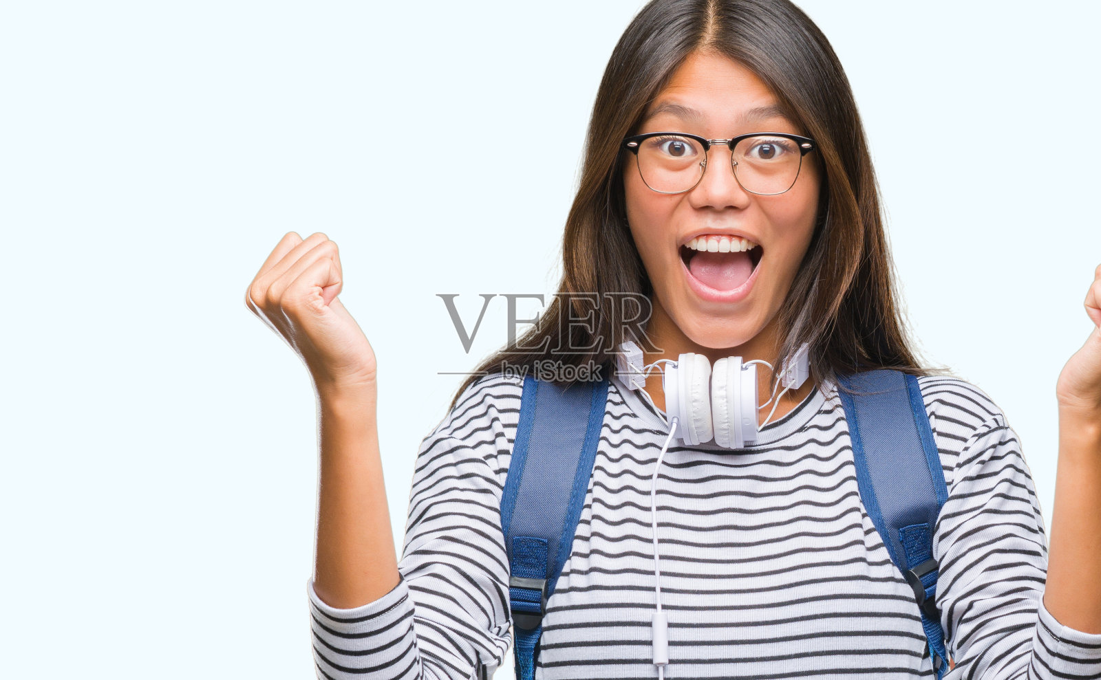 年轻的亚洲女学生戴着耳机和背包,在孤立的背景下举起手臂,睁大眼睛庆祝成功的惊喜和惊奇。赢家的概念。照片摄影图片