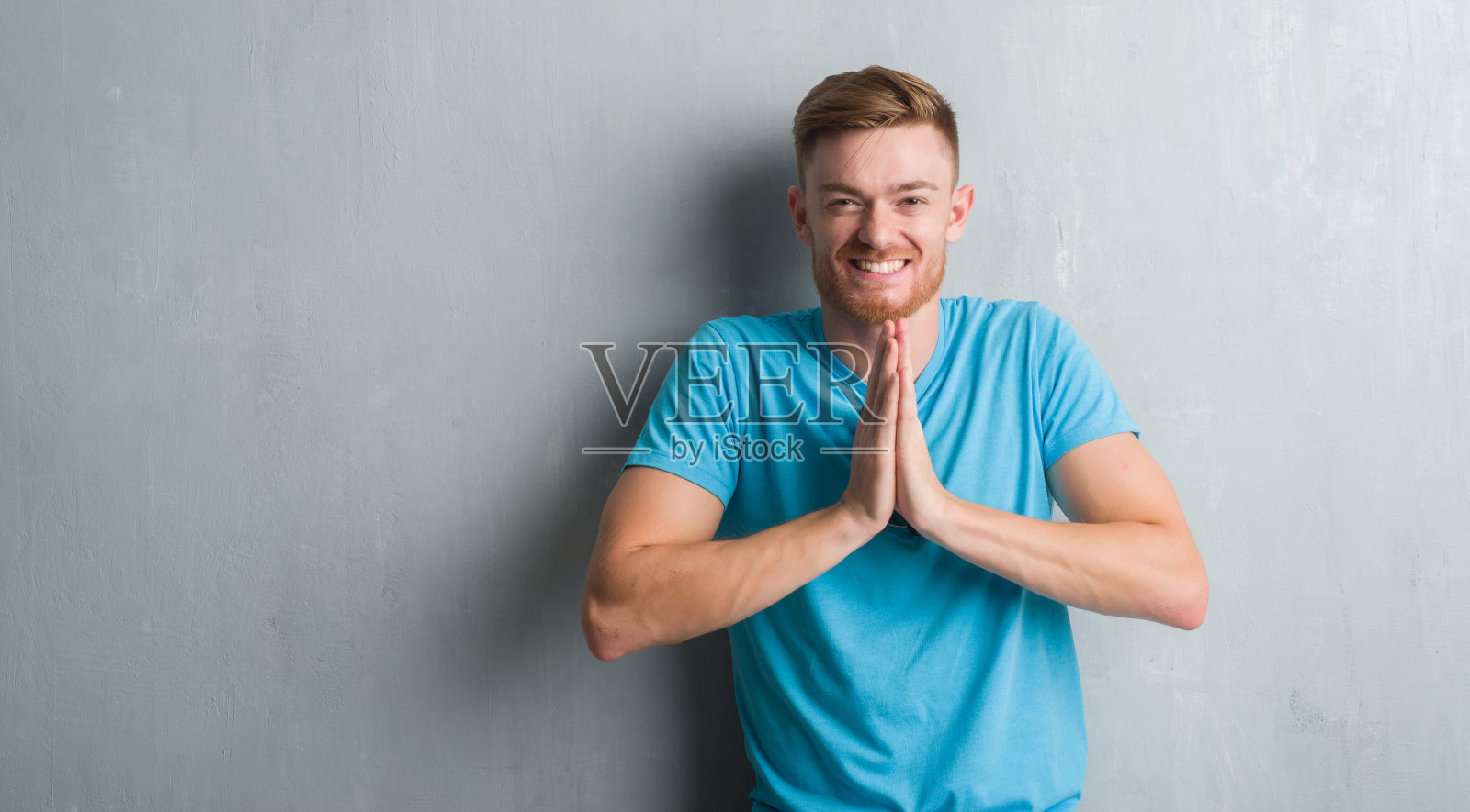 年轻的红发男子在灰色垃圾墙穿着休闲服装祈祷与双手一起请求原谅自信的微笑。照片摄影图片