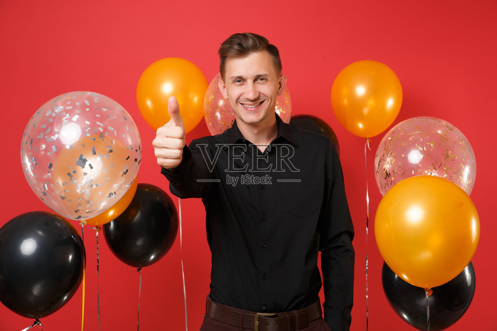 快乐的年轻人穿着黑色经典衬衫庆祝,在红色背景的气球上竖起大拇指。情人节,国际妇女节,新年快乐,生日模型节日派对概念。照片摄影图片