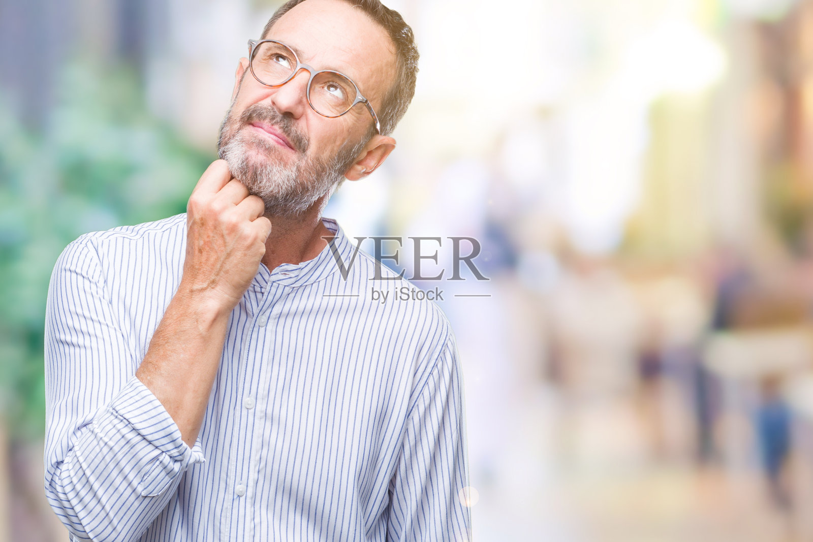 白发苍苍的中年老人戴着眼镜越过孤立的背景,手托着下巴思考问题,神情沉思。带着沉思的微笑。怀疑的概念。照片摄影图片