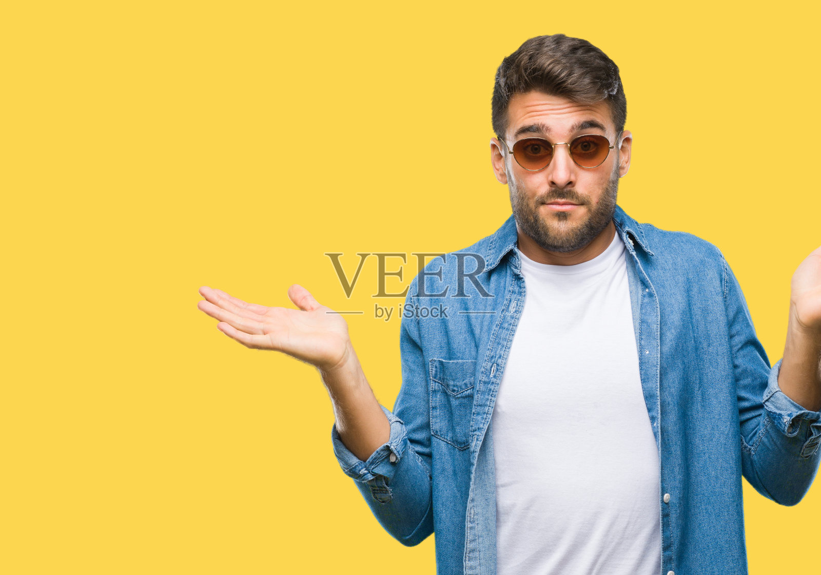 年轻英俊的男子戴着太阳镜在孤立的背景和愚蠢和困惑的表情与胳膊和手举起。怀疑的概念。照片摄影图片