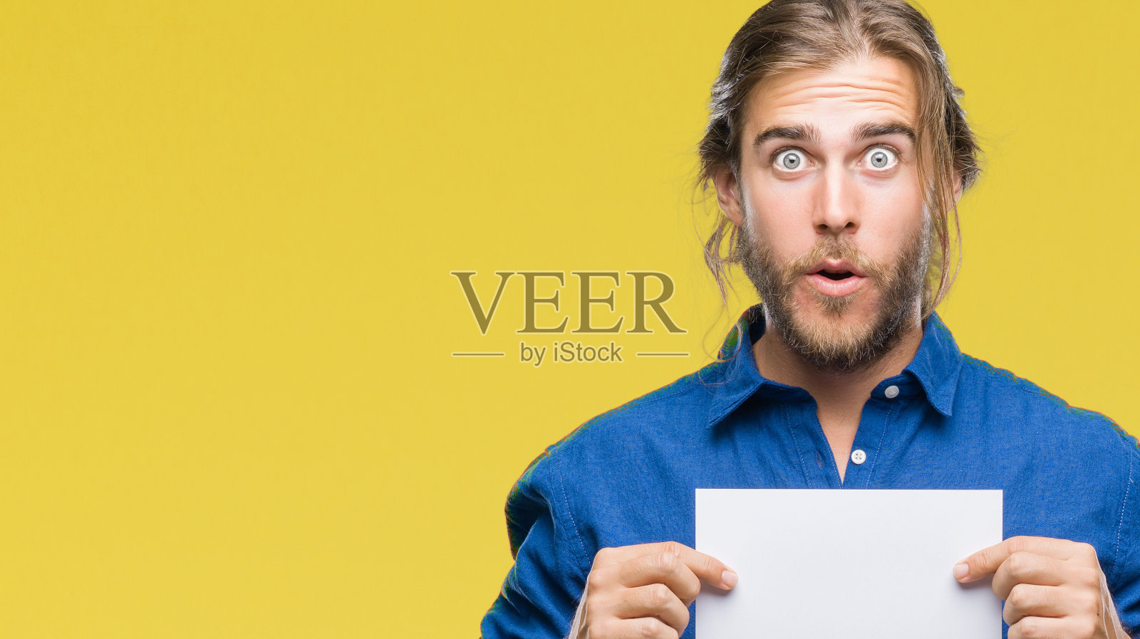 年轻英俊的男子长着长发,隔着孤立的背景,拿着空白的纸吓得目瞪口呆,脸上带着惊恐和兴奋的恐惧表情照片摄影图片