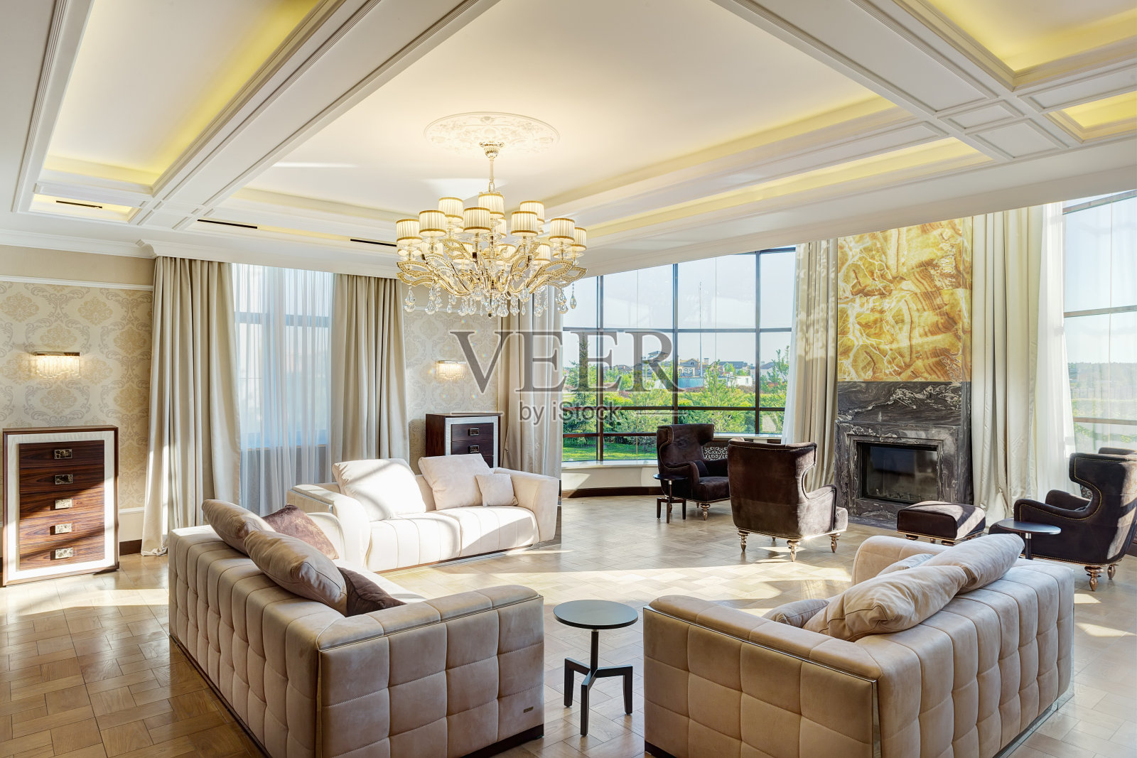 豪华的客厅,大窗户,花园全景和水晶吊灯的中心天花板。室内装饰时尚,壁炉周围有三个沙发和扶手椅。照片摄影图片