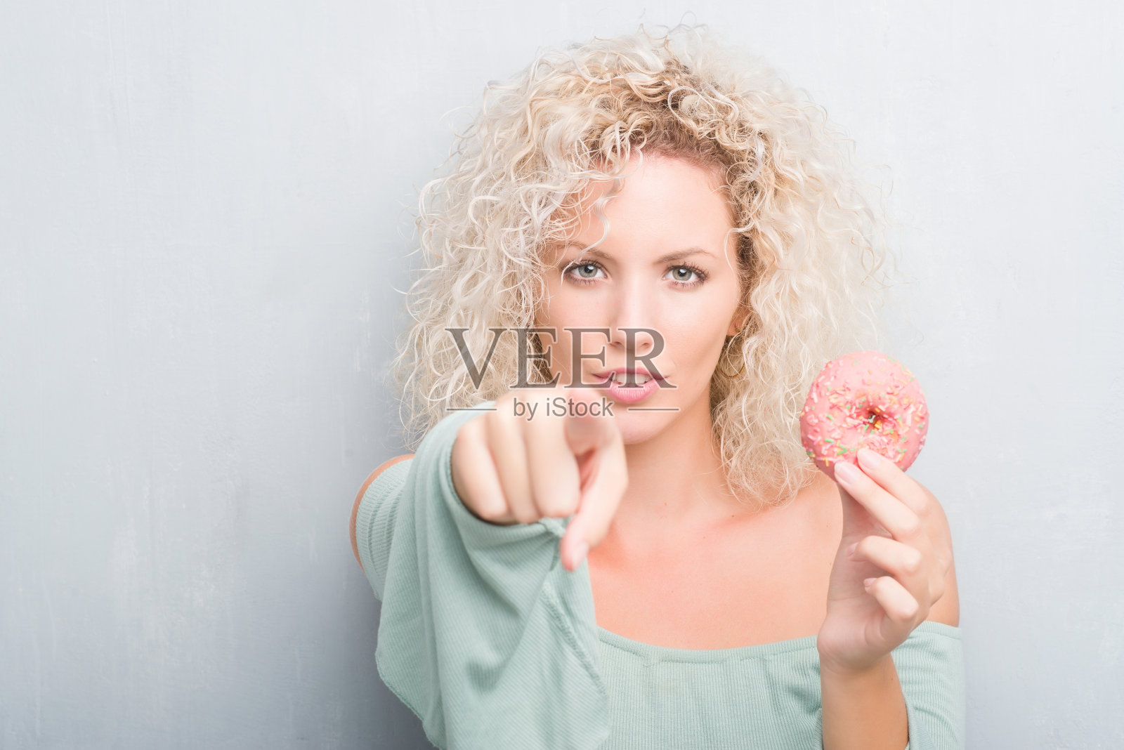 年轻的金发女子在垃圾灰背景吃粉色甜甜圈指向相机和你,手势,积极和自信的手势从前面照片摄影图片