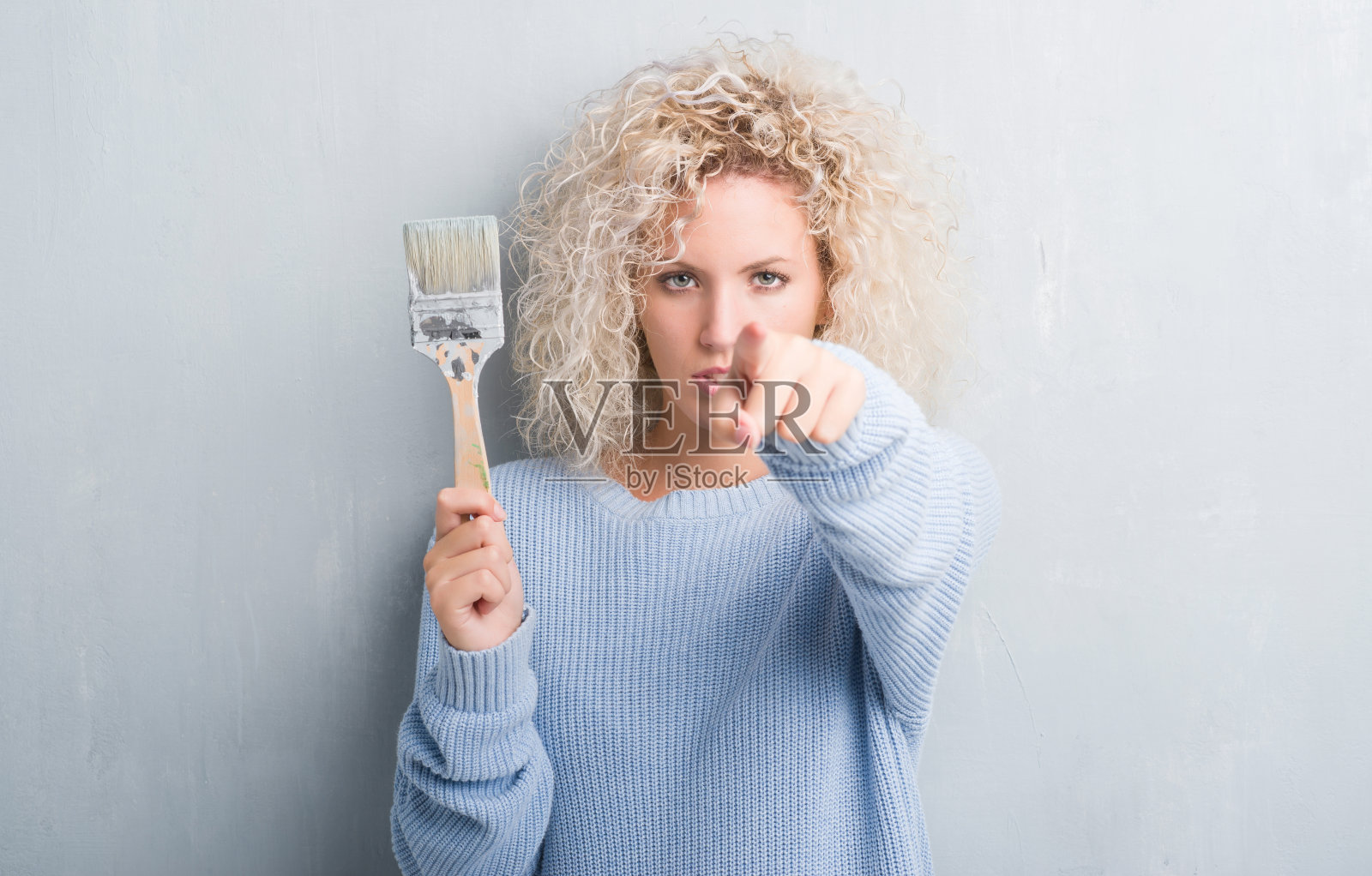 年轻的金发女郎,一头卷发,背景是肮脏的灰色,手里拿着画笔,手指指向相机和你,手势,正面自信的手势照片摄影图片