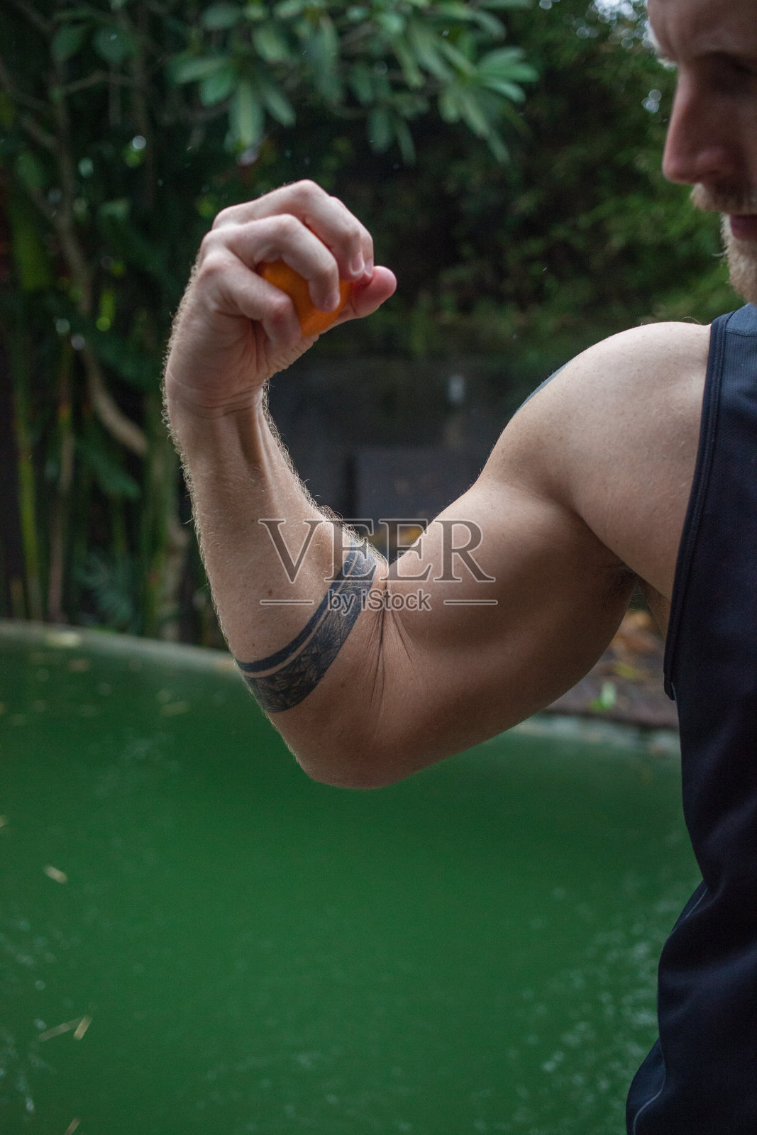 照片中,一名男子在泳池边捏着一个橙子,橙汁在他的手臂上流淌,他身上有纹身,他的肱二头肌和肩膀都很强壮,展示了一种充满水果和蔬菜的健康生活方式照片摄影图片