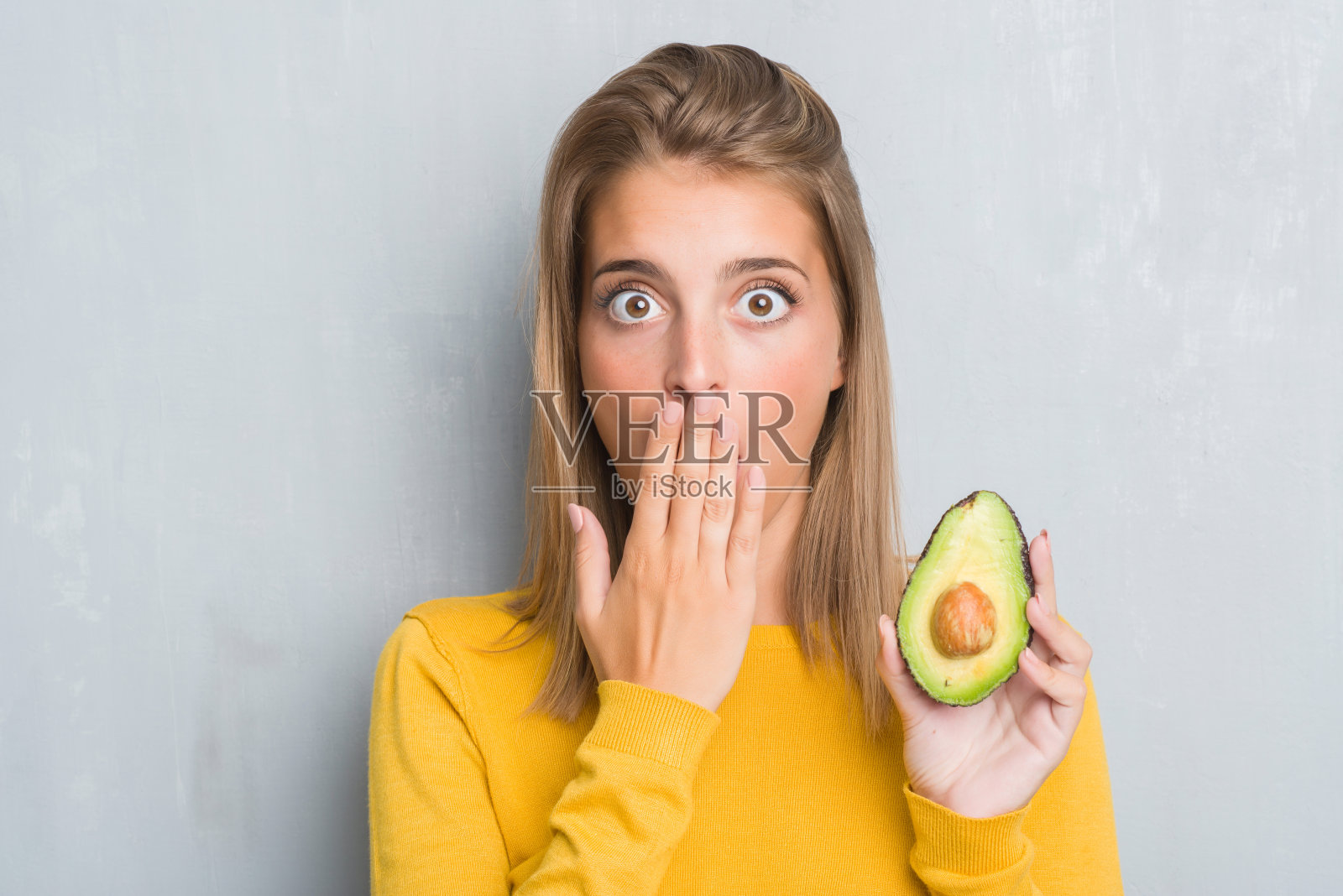 美丽的年轻女子越过脏兮兮的灰墙吃牛油果,捂着嘴用手为错误感到震惊,表情恐惧,在沉默中害怕,秘密的概念照片摄影图片
