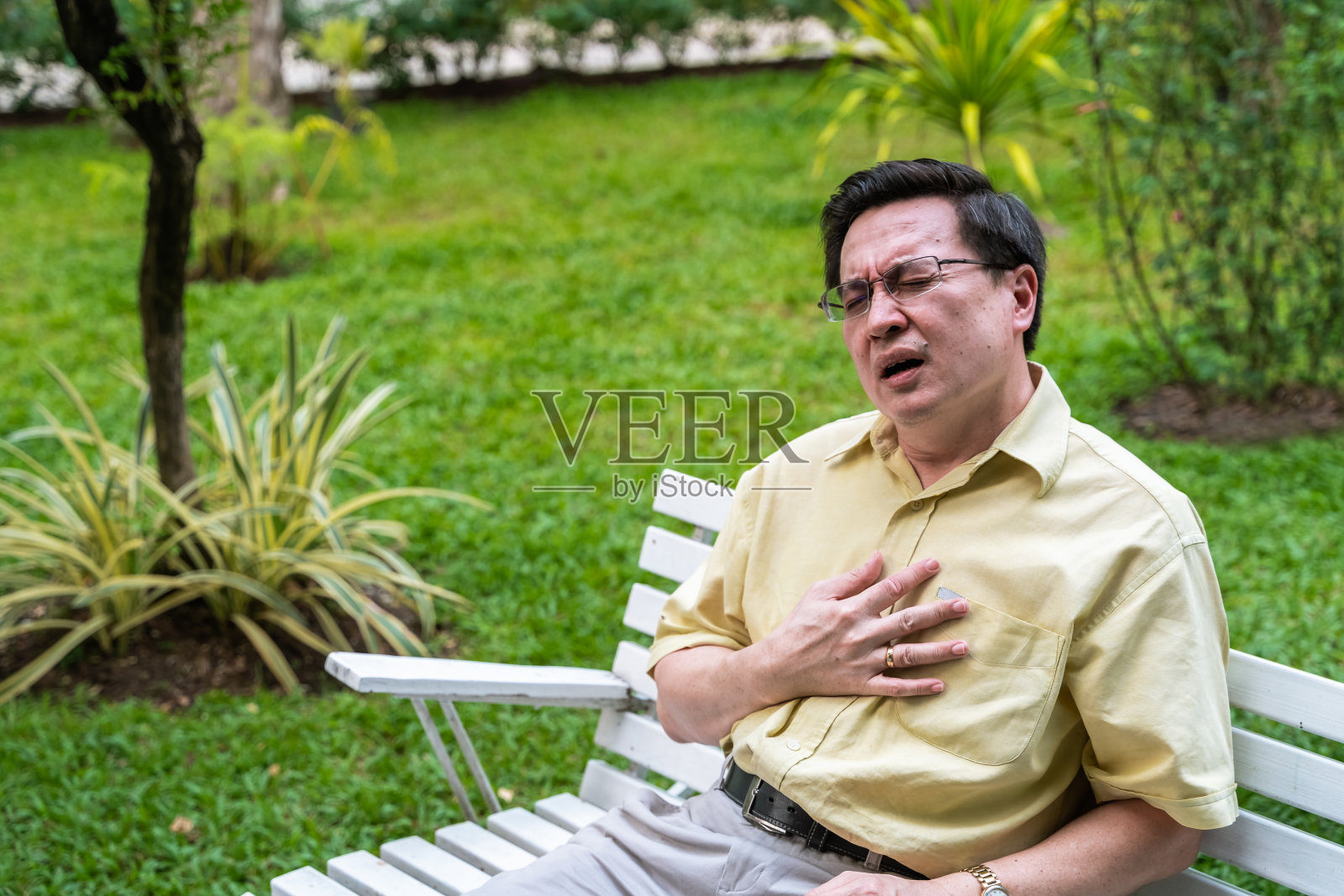 心脏病,亚洲老人抱着胸口,感觉着心脏病发作的痛苦,坐在自家花园的户外扶手椅上。照片摄影图片