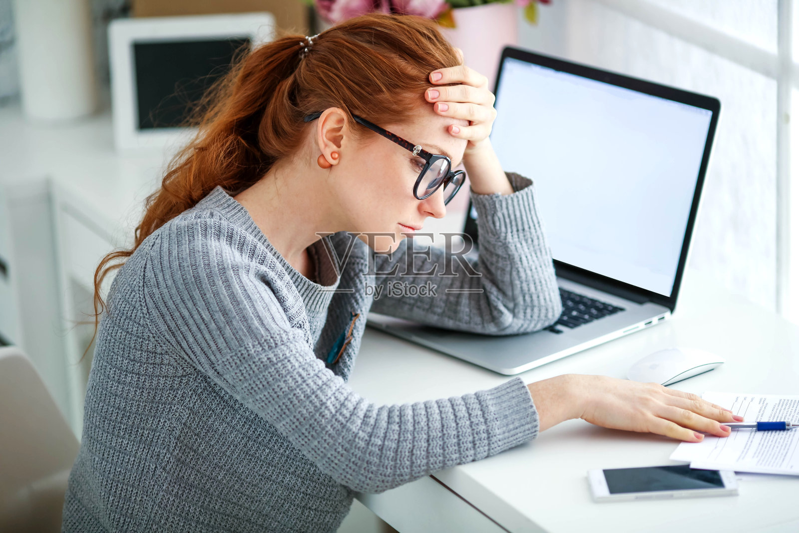 疲惫的红发、戴眼镜的上班族,在办公室工作,使用笔记本电脑和手机照片摄影图片