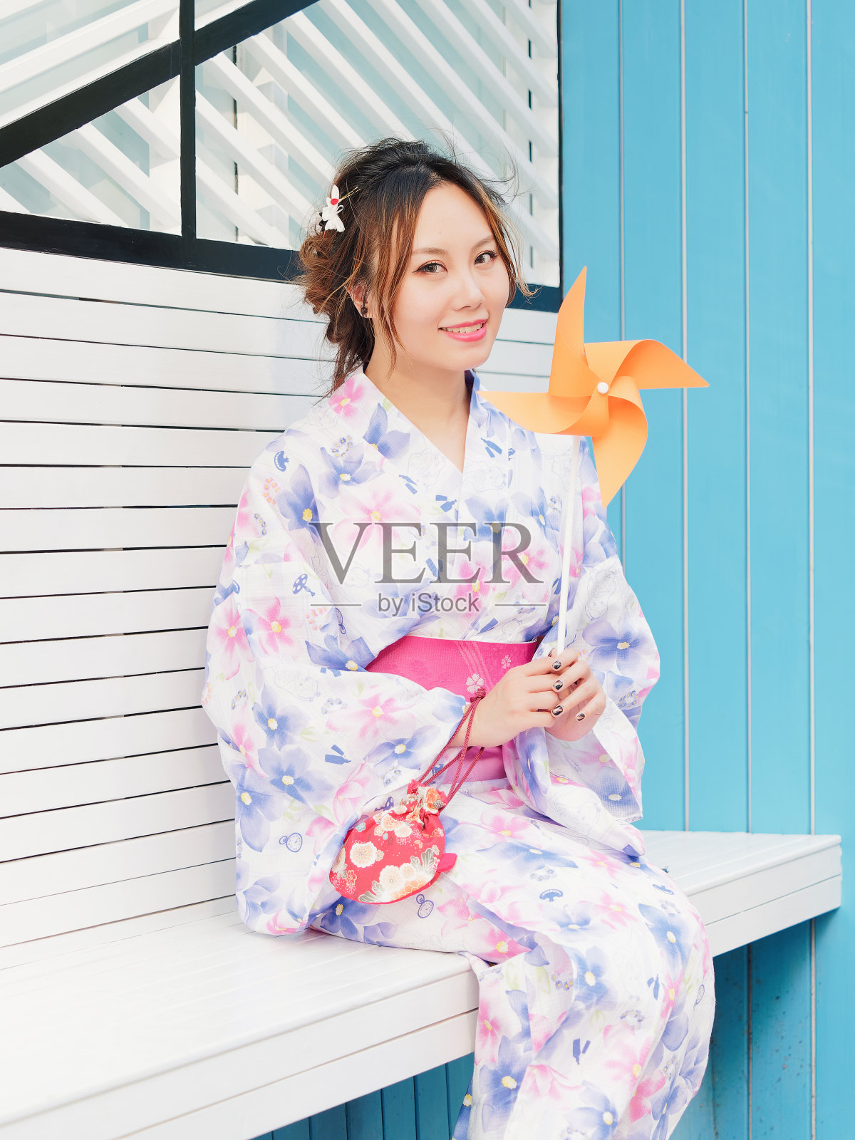 在阳光明媚的日子里,穿着传统日本和服,坐在白色的长凳上玩着她的纸风车的迷人的亚洲女人的肖像,真实的人的生活方式。照片摄影图片