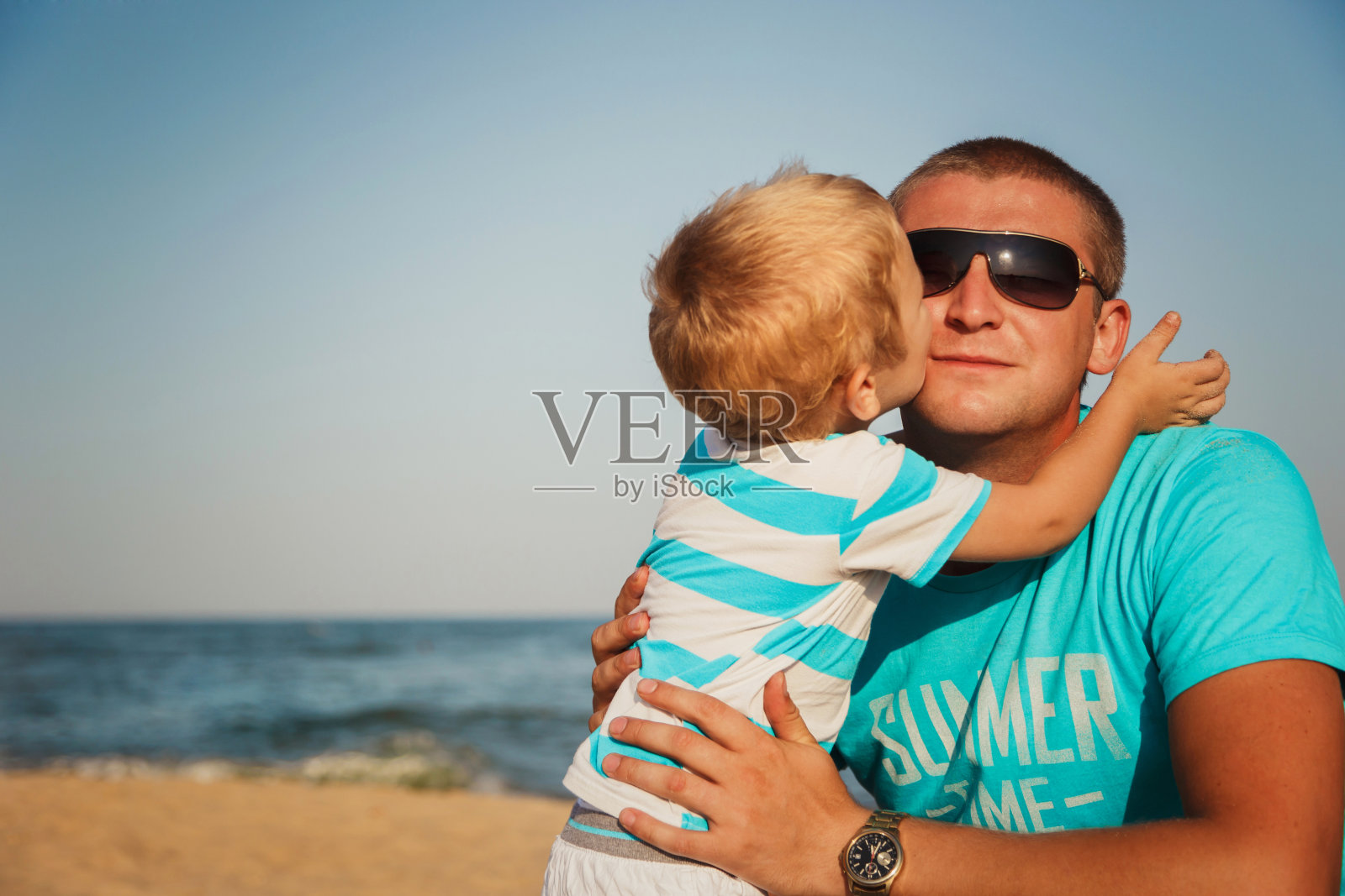 快乐的父子俩在海滩相聚。儿子正在吻他的父亲照片摄影图片