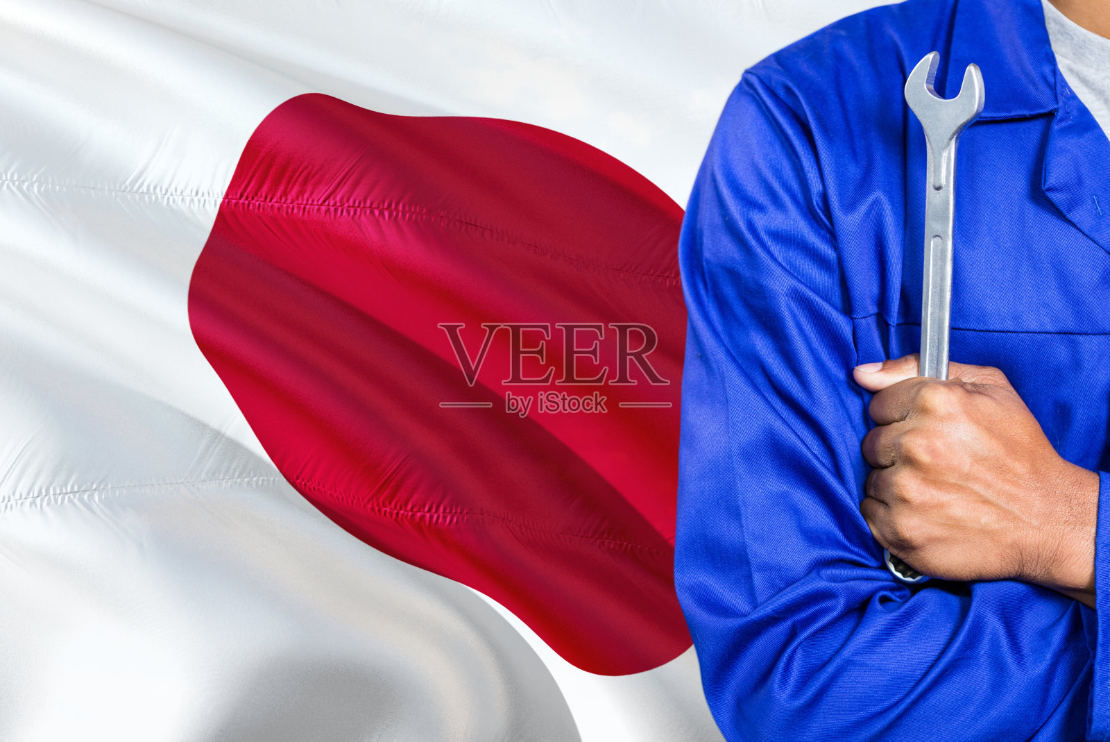 身穿蓝色制服的日本机械师正拿着扳手对抗挥舞的日本国旗背景。交叉双臂技师。照片摄影图片