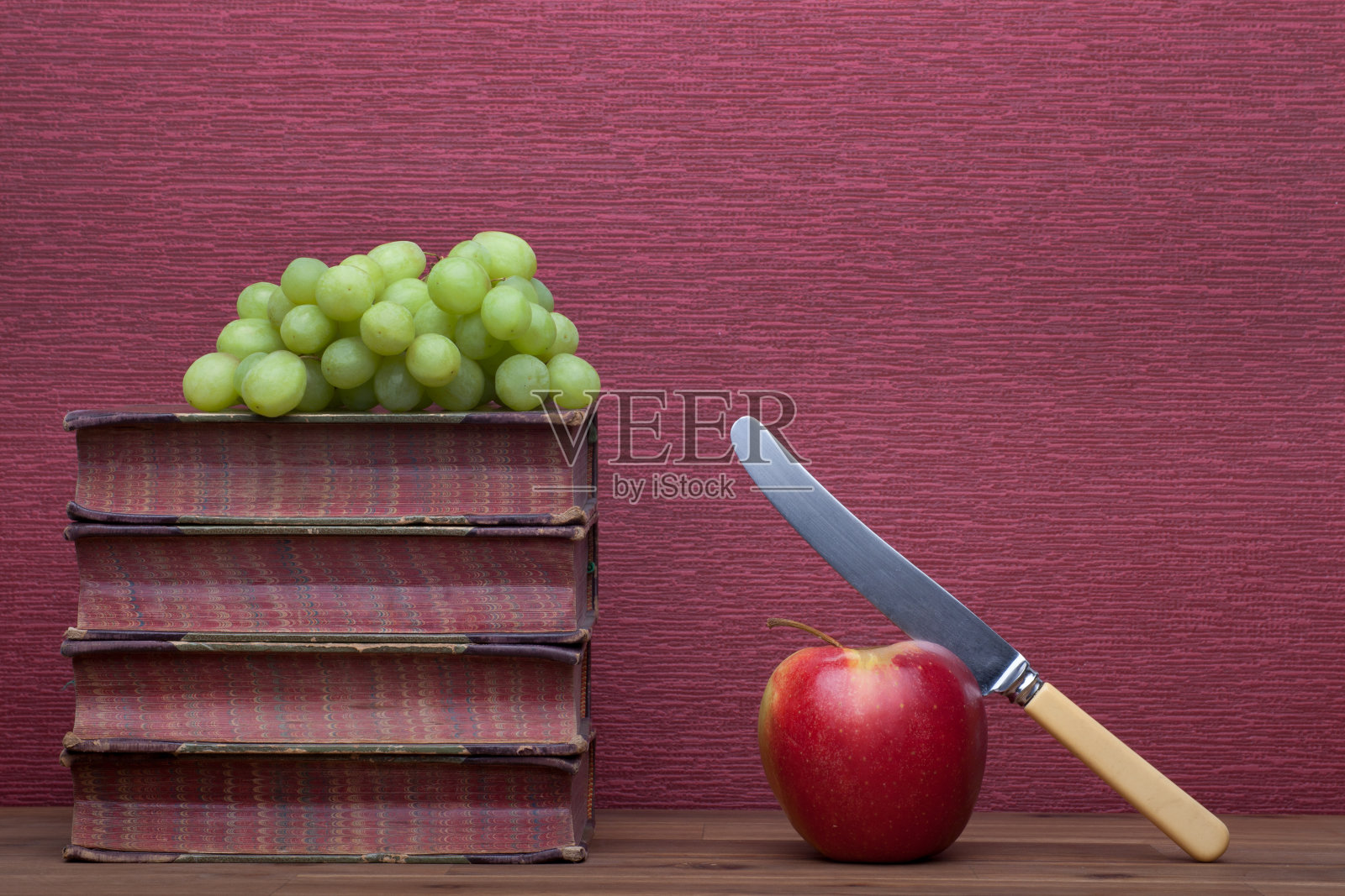 旧书,带刀的苹果,紫红色背景上的葡萄。照片摄影图片